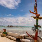 Un colorido cartel de madera en una playa de arena señala "Haynes Cay" y "Paradise", con aguas turquesas transparentes, gente nadando y una estructura con tejado de paja a lo lejos bajo un cielo azul con nubes.