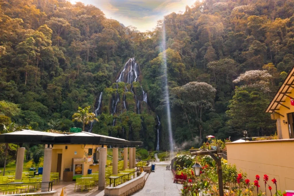 Una alta cascada cae por una exuberante ladera verde detrás de un patio con asientos al aire libre en Termales Santa Rosa, rodeado de árboles y plantas en flor bajo un cielo parcialmente nublado.