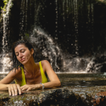 Una mujer con bañador amarillo se relaja en una piscina natural de Termales Santa Rosa, apoyada en una roca, con los ojos cerrados y el pelo mojado hacia atrás. El agua cae en cascada en el fondo, creando una atmósfera serena.