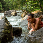 Una mujer con un vestido rosa se agacha junto a un río en Termales Santa Rosa, ahuecando y salpicándose agua en la cara entre grandes rocas y frondosos árboles verdes.