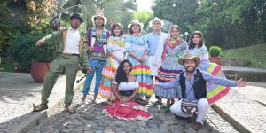 Un grupo de personas ataviadas con coloridos trajes tradicionales posa al aire libre en un camino empedrado del Parque Consotá. La mayoría están de pie y sonríen, mientras que una mujer se arrodilla en el centro, rodeada de frondosos árboles y plantas.
