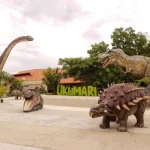 Estatuas de dinosaurios de tamaño natural, entre ellas un Brachiosaurus y un Tyrannosaurus rex, expuestas al aire libre junto a zonas verdes y un edificio con tejado naranja en el Bioparque Ukumarí de Pereira (Colombia). Al fondo se ve el cartel de Ukumarí.