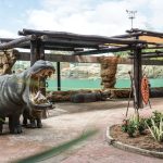 Una exposición zoológica en el Bio-parque Ukumarí presenta una gran estatua de hipopótamo con la boca abierta, carteles informativos, plantas exuberantes, un banco y dos figuras de hipopótamo a tamaño real detrás de una barrera bajo un cielo parcialmente nublado.