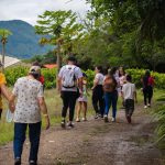 Un grupo de personas, entre las que hay adultos y niños, camina por un sendero de tierra en el Parque de la Uva, un exuberante entorno verde al aire libre con árboles y montañas al fondo. Algunos llevan mochilas y van cogidos de la mano.