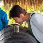 En el Parque de la Uva, una mujer se inclina hacia delante para oler la abertura de un gran barril de madera, mientras un hombre con camisa azul permanece cerca bajo un tejado de paja. Al fondo se ve una exuberante vegetación.