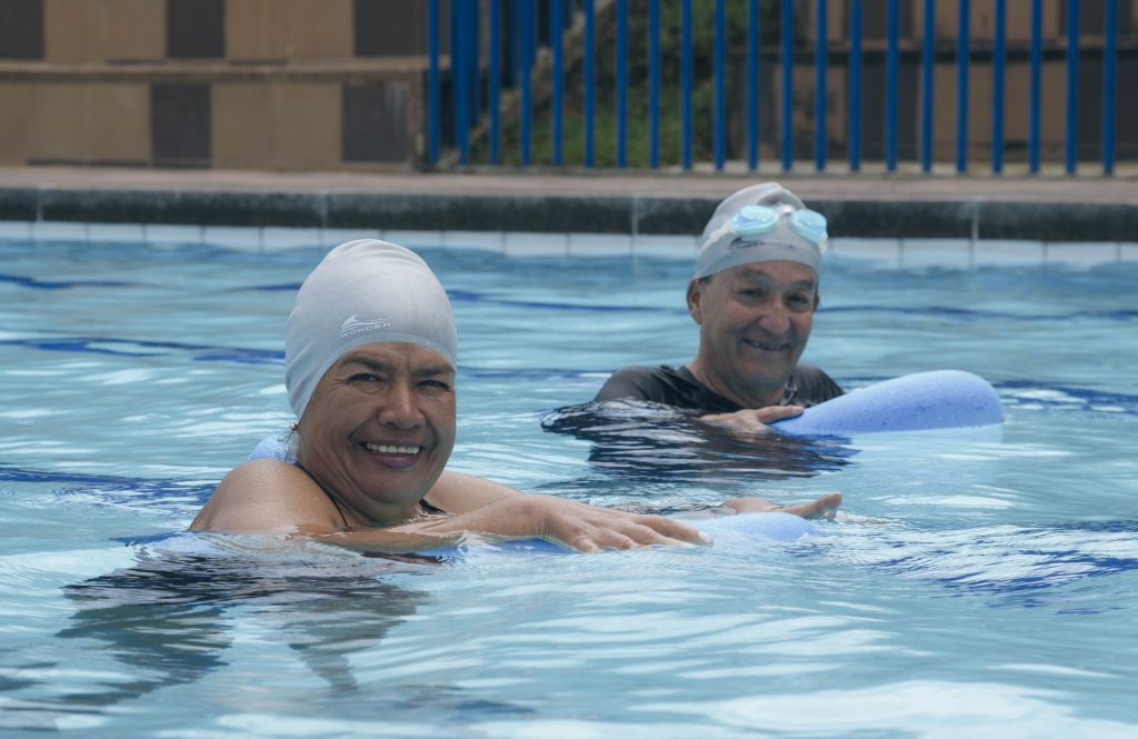 Dos personas mayores con gorros y gafas de natación sonríen mientras sostienen tablas de patada en la piscina, disfrutando su tiempo en el agua.