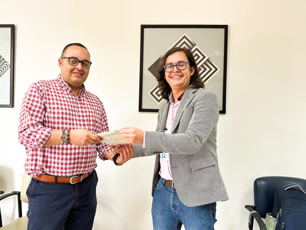 Dos personas sonríen mientras una entrega un documento a la otra en una oficina. Ambos llevan gafas y un atuendo informal de negocios, mientras que en el fondo se ve arte geométrico enmarcado y una insignia de Nosotros UID.