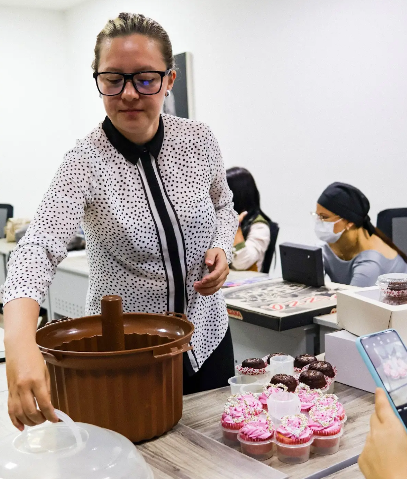Una mujer con gafas y camisa de lunares, que forma parte de una ruta de emprendimiento, destapa productos horneados sobre una mesa, entre ellos magdalenas con glaseado rosa. Cerca, otros se sientan juntos: uno lleva una máscara y otro sostiene un teléfono.