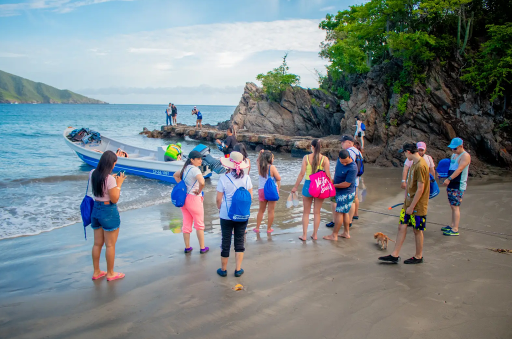 Un grupo de personas con mochilas se encuentra en una playa de arena cerca de una zona rocosa y una pequeña embarcación, disfrutando de un día soleado junto al mar. Algunos, que hablan de Gestión Empresarial, hacen fotos mientras otros admiran el agua y el entorno.
