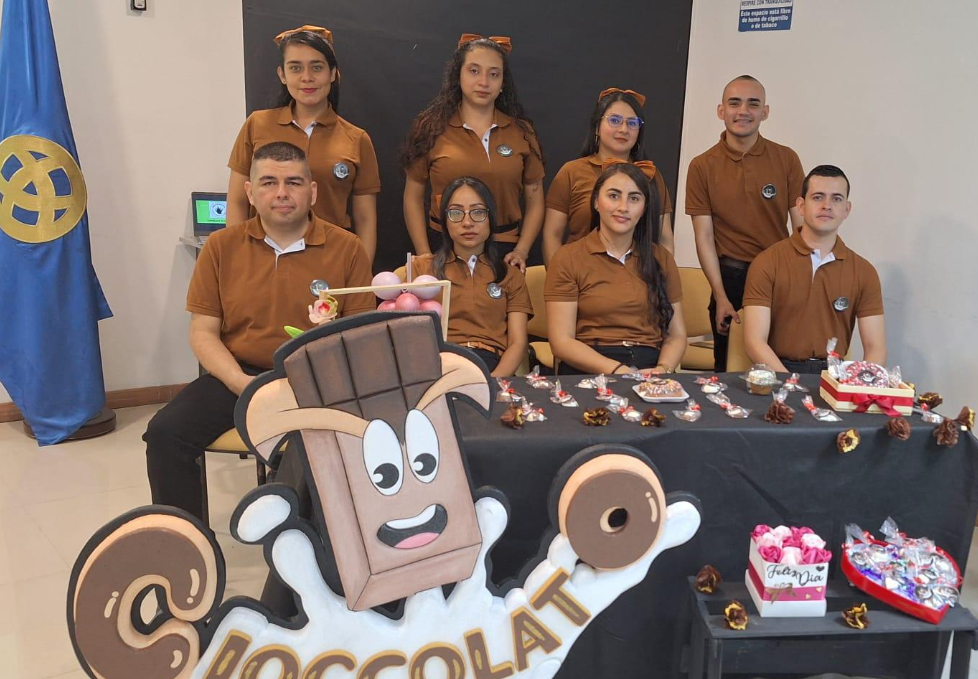 Ocho personas con camisas marrones a juego posan detrás de una mesa en la que se exponen dulces y regalos con temática de chocolate, que representan vivencias empresariales del Instituto de Capacitación Comfamiliar, con una mascota de chocolate de dibujos animados recortada y una bandera azul de fondo.