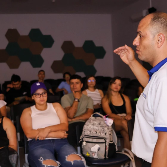 Un hombre con un polo blanco habla a un grupo sentado en un aula sobre cómo construir un hogar. La mayoría de los asistentes parecen atentos, algunos con los brazos cruzados. Una mochila gris con un gato de dibujos animados está sobre una silla en primer plano.