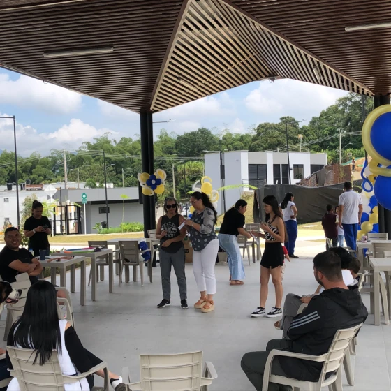 La gente se reúne bajo un pabellón decorado con globos azules y amarillos. Varias mujeres conversan de pie en el centro mientras otras se sientan en sillas de plástico, disfrutando de la vista de casas, vegetación y un cielo parcialmente nublado al fondo.