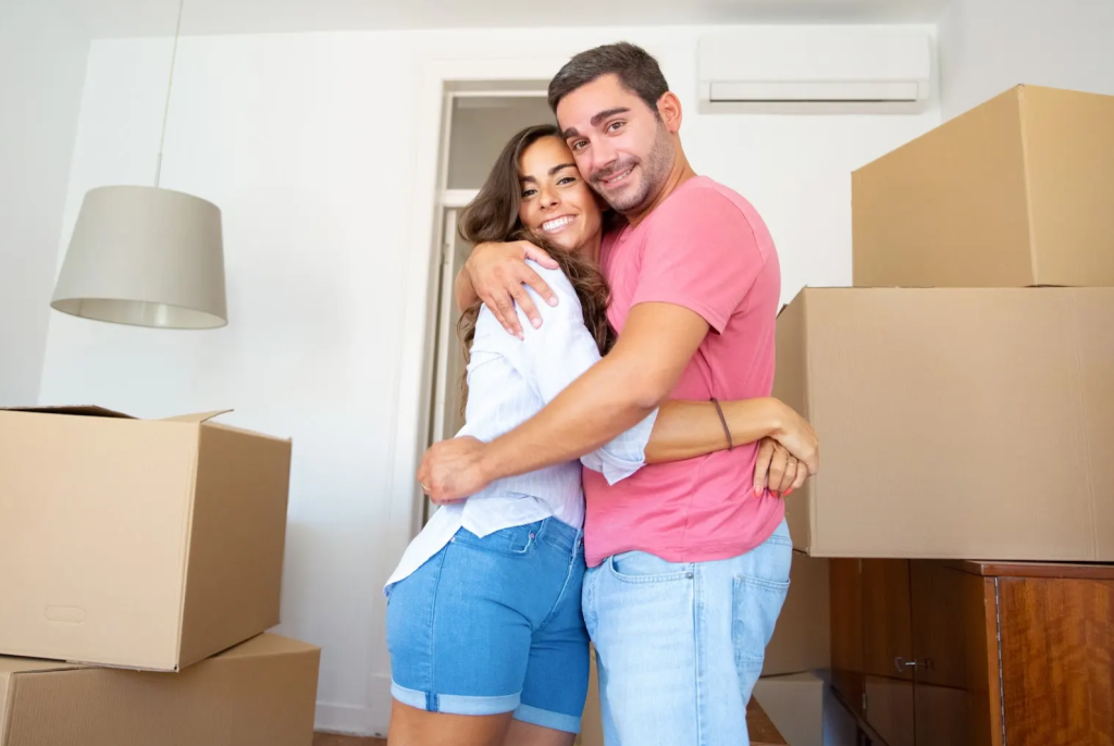 Una pareja sonriente se abraza en una habitación llena de grandes cajas de cartón, lo que sugiere que se están mudando a un nuevo hogar. Ambos parecen felices y relajados, de pie, muy juntos entre las cajas desembaladas.
