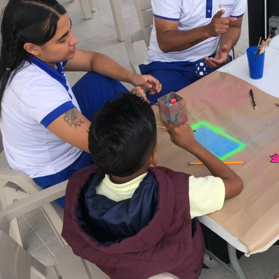 Un niño se sienta en una mesa con dos adultos, trabajando en un colorido proyecto de arte sobre la construcción de hogares. Pinturas, pinceles y una pequeña caja de cartón se reparten mientras crean juntos la casa de sus sueños.