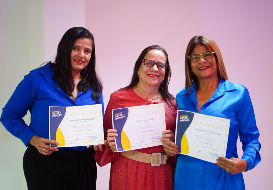 Tres mujeres sonríen y sostienen certificados, de pie, una al lado de la otra, sobre un fondo rosa y blanco. Para celebrar su emprendimiento, dos mujeres visten de azul y una de rojo.