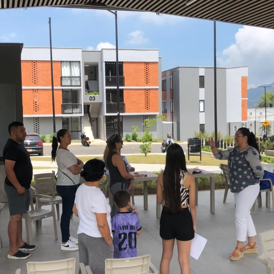 Un grupo de personas, entre las que hay adultos y niños, escuchan a una mujer que habla al aire libre, bajo una zona cubierta. Al fondo se ven edificios de apartamentos, que aluden a la singular historia de construcción de cada hogar, y un coche aparcado.