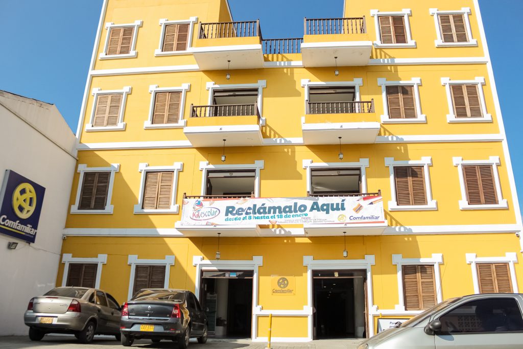 Un edificio amarillo brillante de tres plantas con contraventanas y balcones marrones exhibe una pancarta en la que se lee "Reclámalo aquí". Ideal para la gestión de empresas, se encuentra bajo el sol, con varios coches aparcados frente a la entrada.