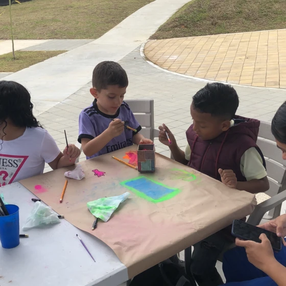 Cuatro niños se sientan alrededor de una mesa al aire libre, pintando y haciendo manualidades con pinturas y pinceles de colores. Mientras se ponen creativos, están imaginando y construyendo tu hogar a su manera artística. La hierba y el pavimento se ven al fondo.