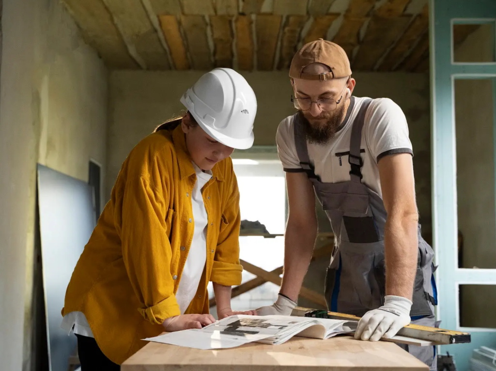 Dos obreros de la construcción, uno con casco blanco y camisa amarilla, el otro con mono y guantes, están de pie ante una mesa mirando planos en una habitación parcialmente renovada.