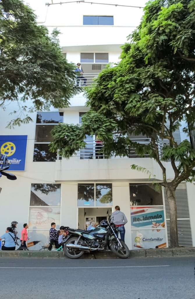 Vista desde la calle de un edificio blanco de tres plantas de Gestión Empresarial con árboles delante, motocicletas aparcadas junto a la acera y gente de pie cerca de la entrada y mirando desde los balcones. En el edificio se ven señales y carteles.