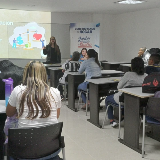 Una mujer presenta una diapositiva en español a una clase de adultos sentados en pupitres, algunos tomando notas; un cartel en el fondo reza "Juntos Construyendo tu Hogar", haciendo hincapié en las ideas sobre construcción y vivienda.