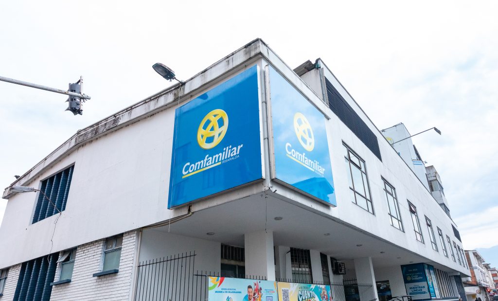 Un edificio blanco con un gran cartel azul con el logotipo amarillo y "Comfamiliar Gestión Empresarial" en la esquina. El cielo está nublado y hay una farola y una cámara de seguridad cerca.