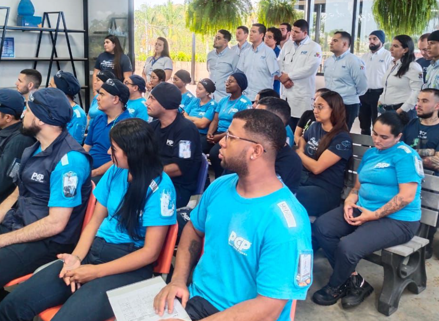 Un grupo de personas vestidas con uniformes azules y grises asisten a una reunión o sesión de formación de Comfamiliar Formación Técnica en Logística; algunas toman notas, mientras unas plantas cuelgan al fondo.