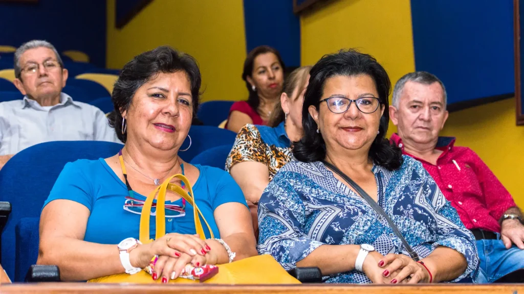 Un grupo de adultos mayores, hombres y mujeres, están sentados en sillas azules en un auditorio, sonriendo y mirando hacia la cámara. Las paredes amarillas y azules de fondo reflejan el espíritu positivo de Aprendiendo a Envejecer.