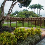 Una pérgola de madera se alza en un exuberante jardín tropical cerca del Auditorio Barranquero, rodeada de arbustos verdes, palmeras y un camino de piedra en un día nublado.