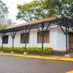Un pequeño edificio blanco con tejado de tejas rojas y contraventanas de madera, rodeado de vegetación y árboles, se alza junto a una carretera de asfalto mojado con el bordillo amarillo, cerca del Auditorio Colibrí.