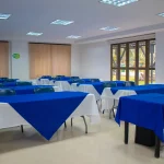 Un aula luminosa del Auditorio Faisán presenta varias mesas cubiertas con manteles azules y blancos, sillas azules, grandes ventanas con persianas y suelo de baldosas. La sala está vacía y bien iluminada con lámparas de techo.