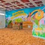 Una colorida sala de fiestas de temática agrícola en el Auditorio Faisán cuenta con balas de heno, virutas de madera en el suelo, decoraciones vegetales y un gran telón de fondo en el que se lee "Bienvenidos Granjeritos" con animales de dibujos animados y un alegre granjero.
