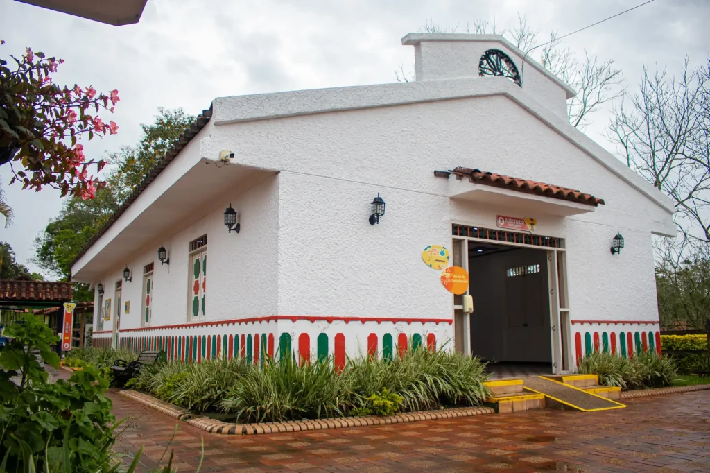 El Auditorio Faisán, un edificio blanco con detalles rojos y verdes, farolillos y tejado de tejas, se asienta sobre un camino de ladrillo rodeado de vegetación. Una rampa amarilla conduce a la entrada bajo un cielo nublado.