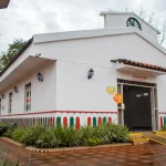 El Auditorio Faisán, un edificio blanco con detalles rojos y verdes, farolillos y tejado de tejas, se asienta sobre un camino de ladrillo rodeado de vegetación. Una rampa amarilla conduce a la entrada bajo un cielo nublado.