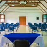 Filas de sillas azules frente a una mesa cubierta con un mantel azul y blanco en el luminoso y espacioso Auditorio Golondrina, con vigas de madera en el techo y grandes ventanales, preparado para una reunión o presentación.