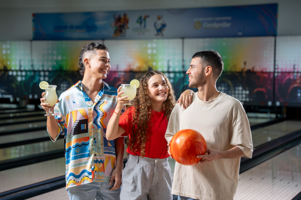 Tres amigos en una bolera sonríen juntos; dos sostienen cócteles con lima y uno sostiene una bola de bolos naranja. En el fondo se ven pistas de bolos y luces de colores.