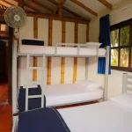 Una luminosa habitación en Cabañas Caracolillo cuenta con una litera y una cama individual, ropa de cama blanca, vigas de madera en el techo, un ventilador de techo, una puerta abierta, una gran ventana con cortinas azules y suelo de baldosas rojas. En el exterior se ven árboles verdes.