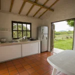Una pequeña cocina rústica en Cabañas Caracolillo cuenta con armarios blancos, un frigorífico y una mesa con sillas blancas. La puerta da a una zona de césped con árboles y vistas a la montaña, mientras que las baldosas de terracota calientan el suelo.