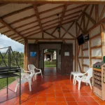 El rústico porche de Cabañas Caracolillo tiene suelo de baldosas rojas, vigas de bambú, sillas blancas de plástico, una parrilla metálica, un zapatero de madera y puertas dobles de madera que dan a una zona exterior cubierta de hierba.