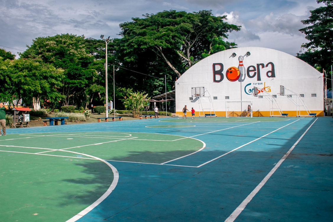 Cancha múltiple al aire libre con marcas verdes y azules, rodeada de árboles. Al fondo, un gran edificio blanco rotulado "Bolera" muestra un gráfico de una bola de bolos y bolos. Hay gente paseando cerca.
