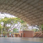 Cancha de baloncesto cubierta en un polideportivo con techo metálico arqueado, gradas vacías a la derecha y árboles visibles en el exterior a través de los laterales abiertos. A la izquierda hay un marcador y una canasta de baloncesto.