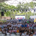 Un grupo numeroso de personas, en su mayoría niños y adolescentes, posan juntos bajo un coliseo polideportivo cubierto con árboles al fondo. Una pantalla detrás de ellos muestra Festival Juntos, mientras en primer plano se disponen sillas blancas.