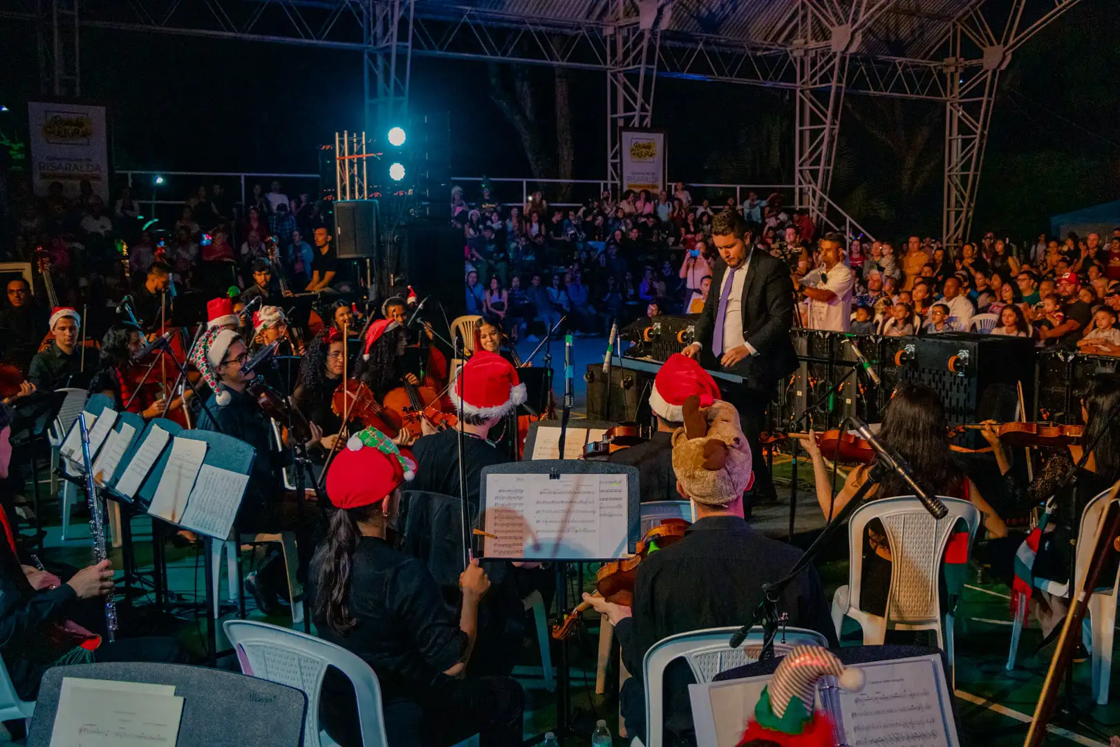 Una orquesta con sombreros de fiesta actúa en el escenario ante el público por la noche. El director se sitúa al frente, y el público se sienta apretujado en un Polideportivo bajo un toldo metálico, disfrutando de la música en este vibrante recinto al aire libre.