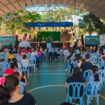 Una gran multitud se sienta en sillas de plástico frente a un escenario en el coliseo cubierto. Personas de todas las edades observan a los artistas bajo el recinto cubierto al aire libre, con árboles visibles al fondo.