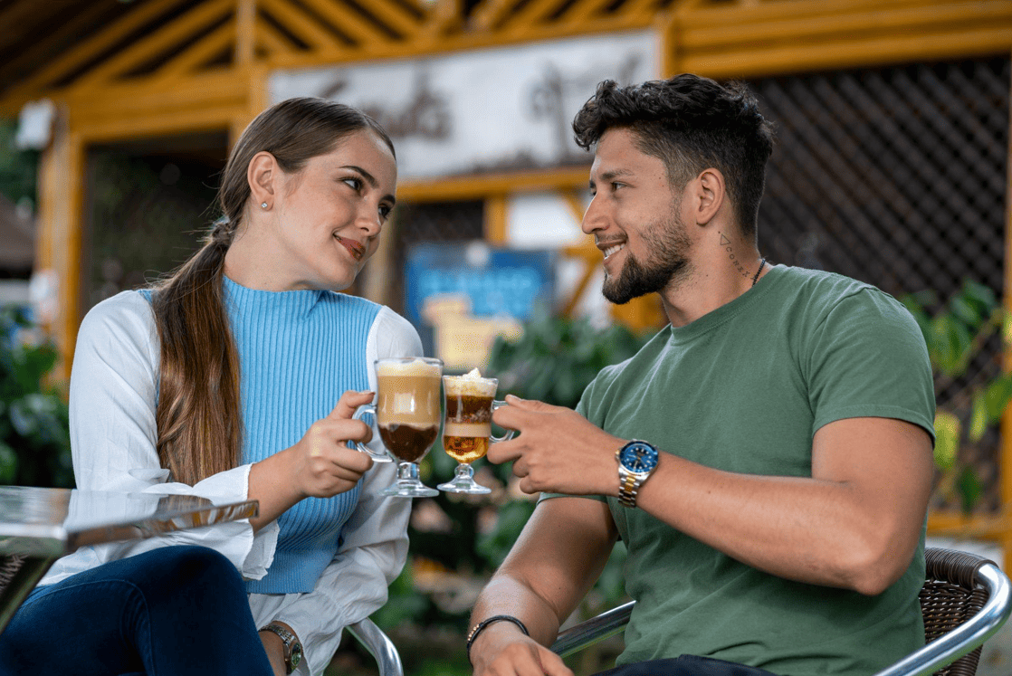 Una mujer y un hombre jóvenes se sientan al aire libre en el café Consotá, sonríen y brindan con sus bebidas. La mujer sostiene un café con leche, el hombre un pequeño vaso de cerveza. Parecen felices y conversan.