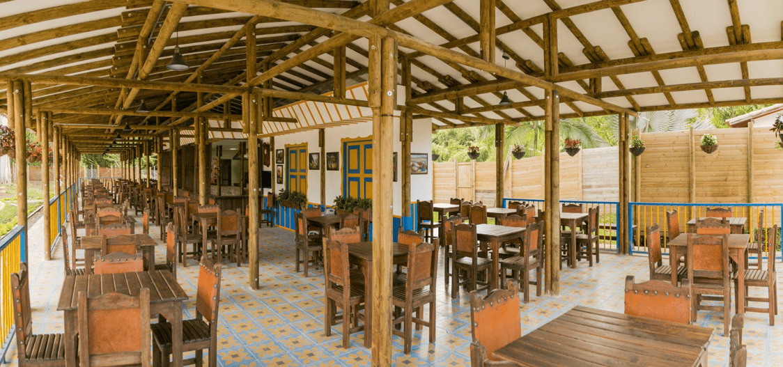Restaurante al aire libre El Montañerito con vigas de madera, suelo de baldosas de colores, mesas y sillas de madera, puertas amarillas y azules, plantas en macetas y una valla al fondo. El espacio parece luminoso y acogedor.