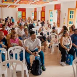 Un grupo heterogéneo de personas se sienta en sillas de plástico blancas en una sala muy iluminada con coloridas puertas y ventanas rojas y amarillas en Pereira Antigua, para asistir a un acto o reunión en el interior.
