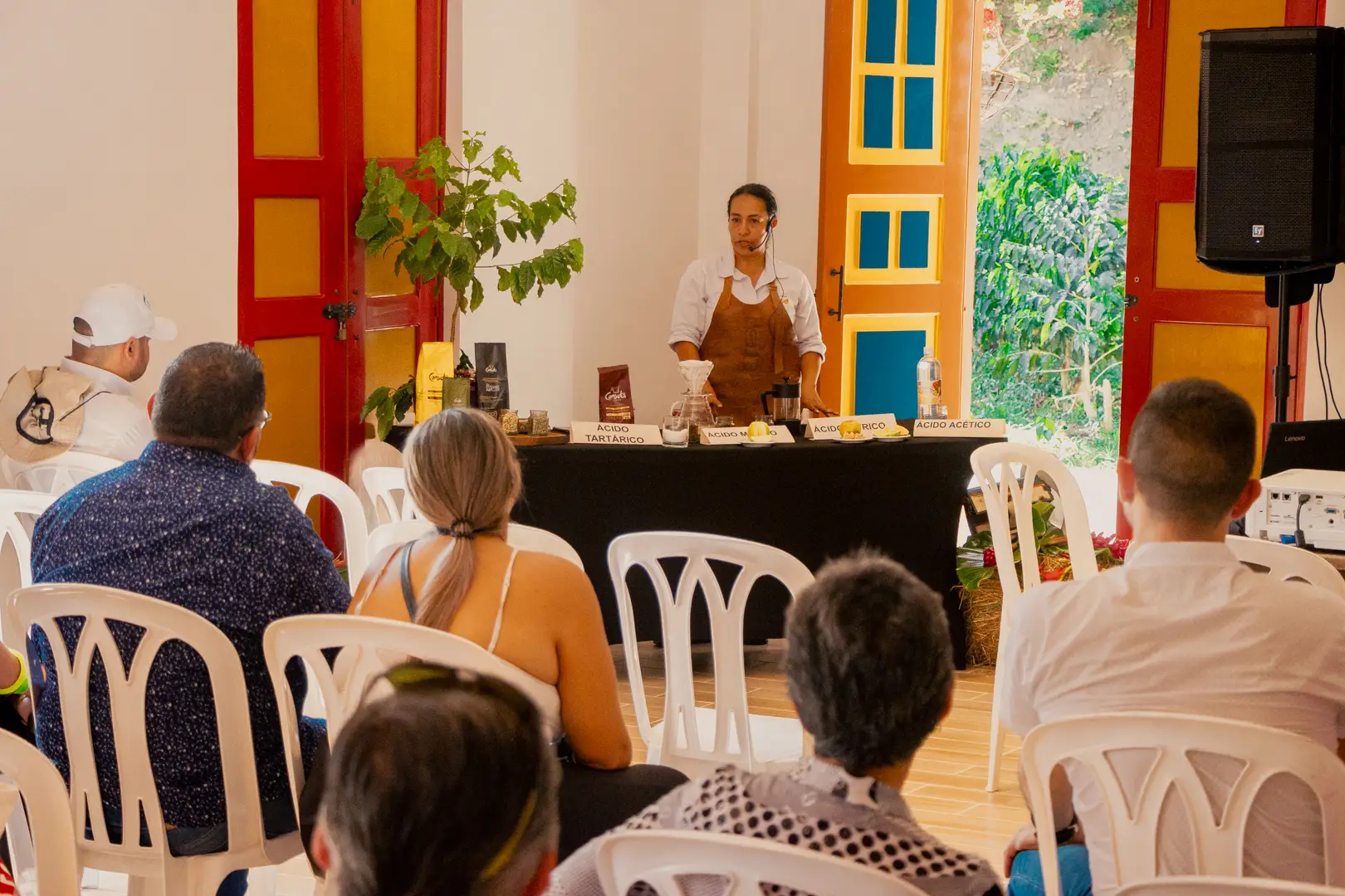 Una persona, de pie detrás de una mesa con varios objetos, habla a un público sentado en sillas blancas en una colorida sala de la Fonda Réplica de Pereira Antigua, con puertas y ventanas de vibrantes colores rojo y amarillo.