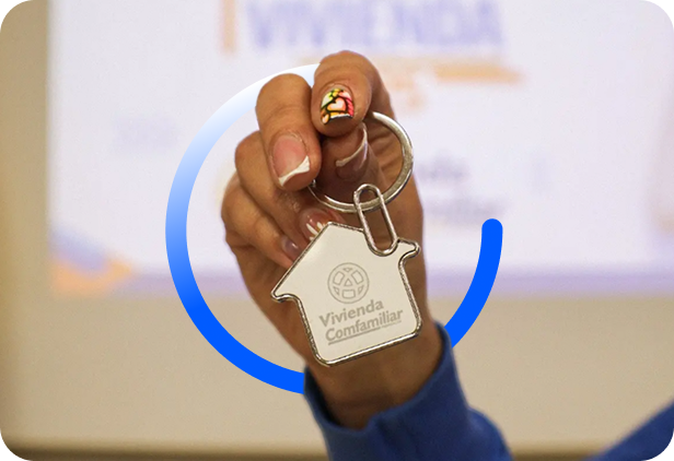 Una mano sostiene un llavero en forma de casa con la etiqueta "Vivienda Confamiliar". El fondo está difuminado y las uñas de la persona están pintadas con un diseño colorido.