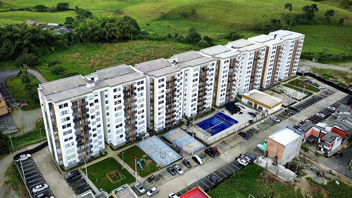 Vista aérea del Mirador Campestre, un moderno complejo de apartamentos con múltiples edificios blancos, una piscina central azul, canchas de baloncesto, zonas de aparcamiento y campos verdes circundantes.